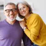 Portrait of happy beautiful senior caucasian family couple in love smiling at camera, retired man and woman hugging embracing while relaxing on sofa in living room at home, enjoying life on retirement