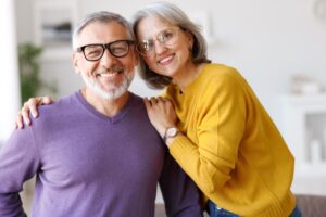 Portrait of happy beautiful senior caucasian family couple in love smiling at camera, retired man and woman hugging embracing while relaxing on sofa in living room at home, enjoying life on retirement